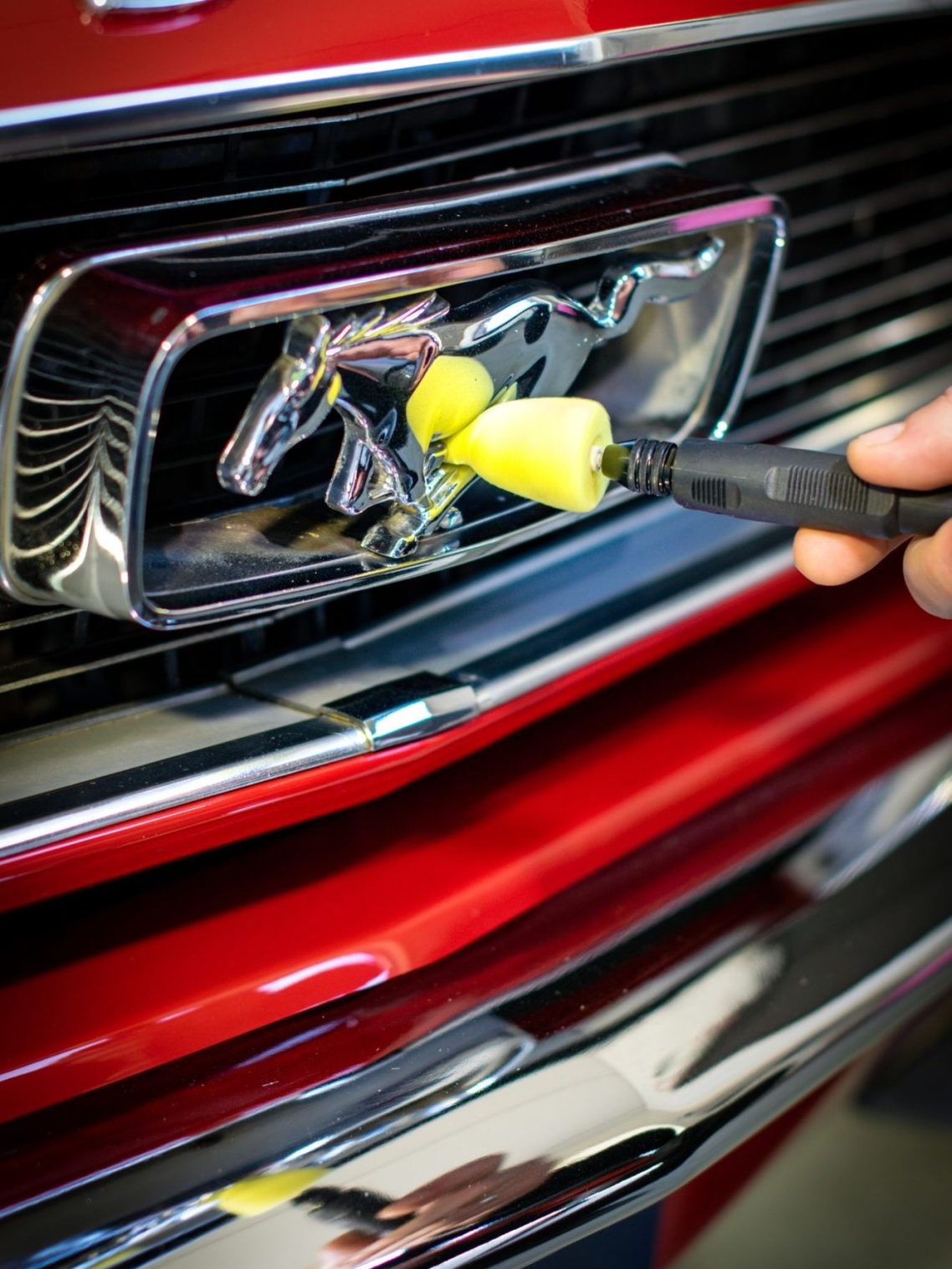 Een hand reinigt een chrome auto-grille met een gele spons op een rode auto.
