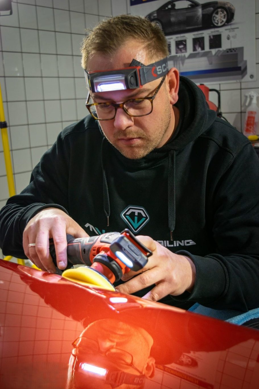 Een man met een hoofdlamp polijst een auto in een werkplaats.